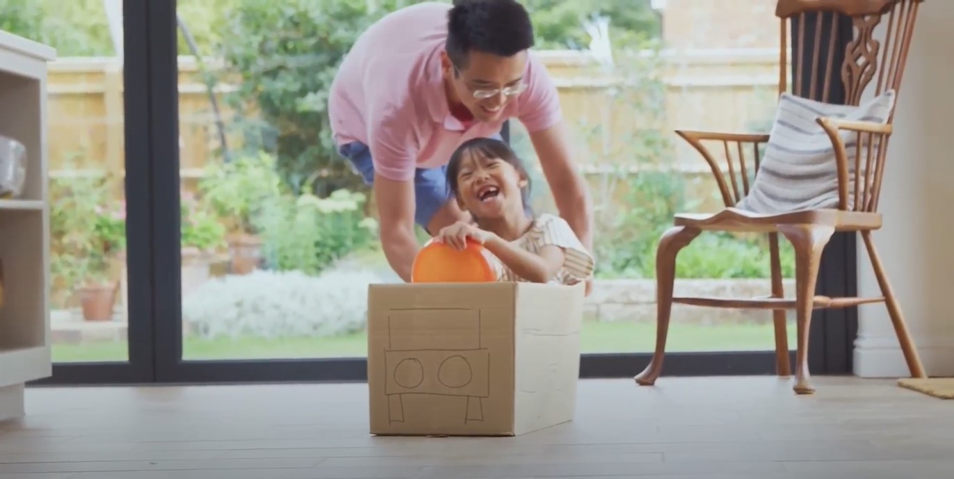 Dad pushing Daughter in Cardboard box on Hardwood Floors in Home.